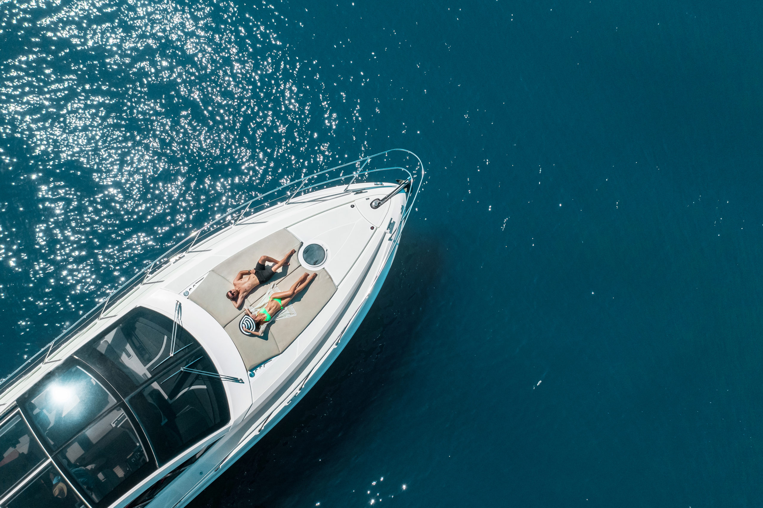 An aerial view of a white luxury yacht floating on deep blue waters, with a person relaxing on the bow deck. Sunlight sparkles across the water's surface, creating a dazzling effect.