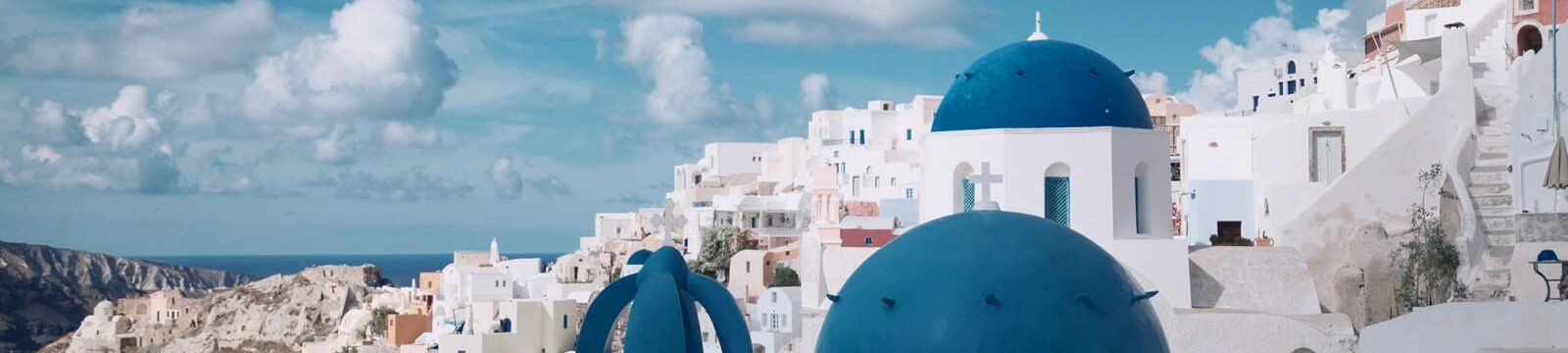 A picturesque view of Oia, Santorini featuring iconic white-washed buildings and distinctive blue-domed churches against a backdrop of blue sky and sea. The traditional Cycladic architecture showcases the island's unique architectural style.
