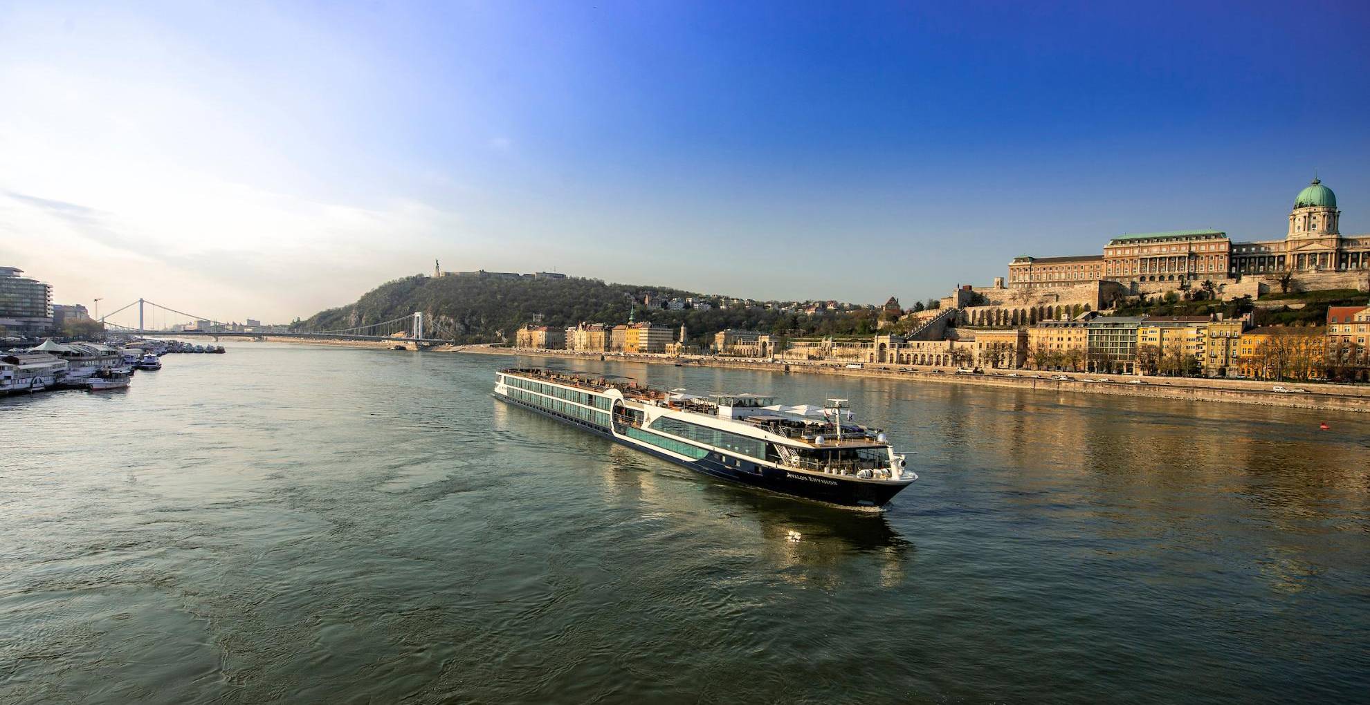 A modern river cruise ship sailing on the Danube River with the historic Buda Castle and Hungarian Parliament buildings in the background. The scenic cityscape showcases Budapest's architectural beauty during golden hour.