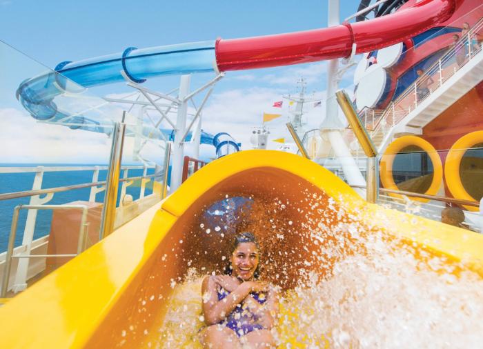 A passenger sliding down a vibrant yellow water slide on a cruise ship, with blue and red water slides in the background and splashing water creating an exciting scene.
