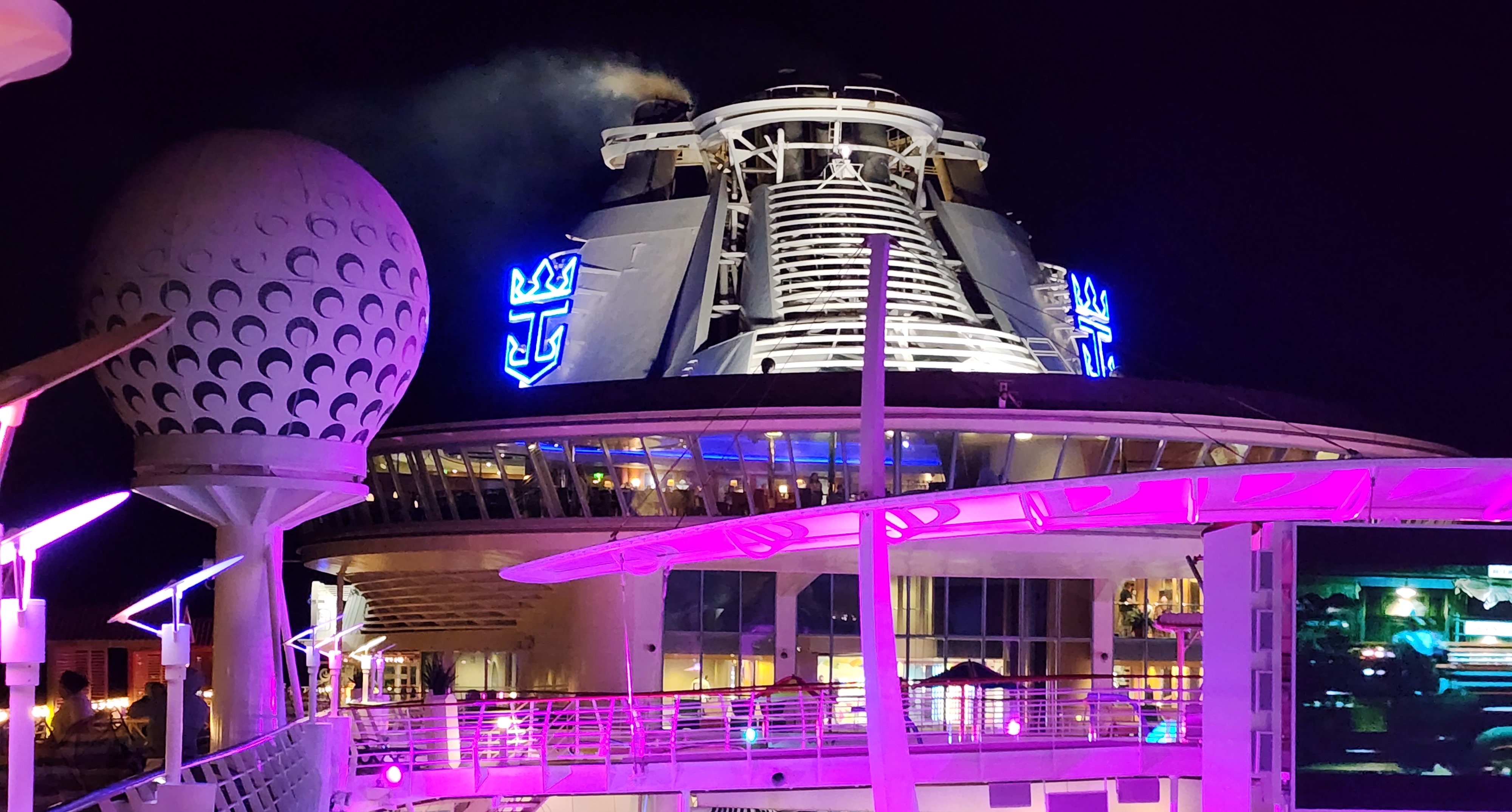 A nighttime view of a Royal Caribbean cruise ship with dramatic purple and blue lighting, showcasing the vessel's modern architecture and illuminated decks.