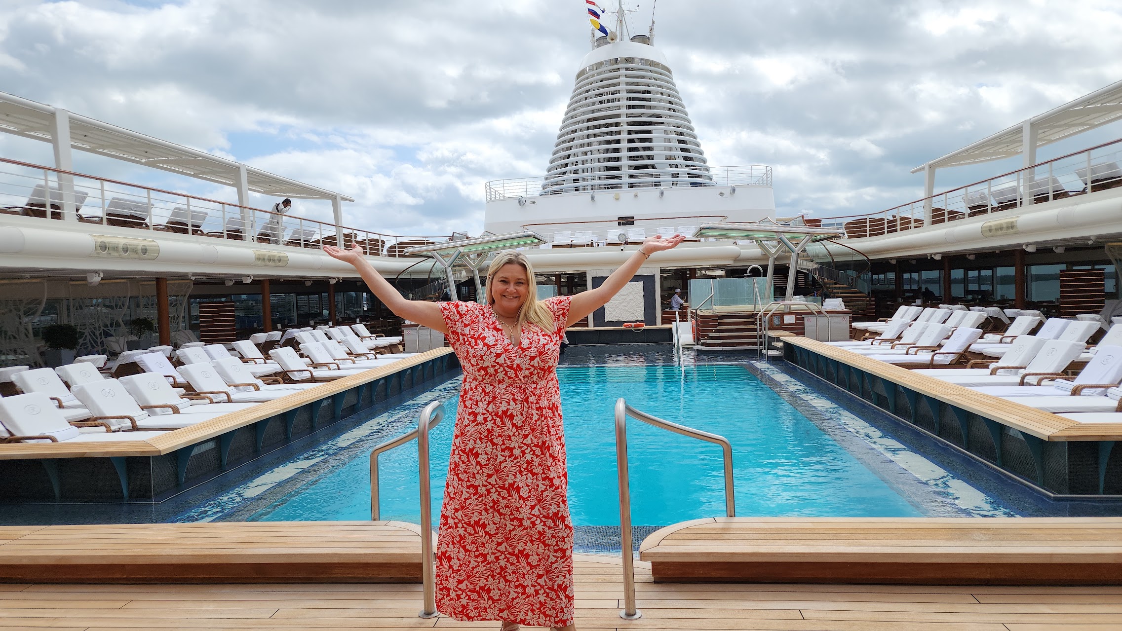 A pristine swimming pool on a luxurious cruise ship with white lounge chairs and a modern design, featuring a smiling passenger in a red floral dress standing with outstretched arms