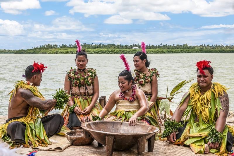 A group of five performers in traditional Polynesian attire sit by a river, preparing and engaging in a cultural ritual involving a large wooden bowl.