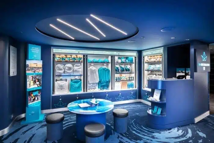 A sleek and modern retail space within a cruise ship, featuring merchandise displays with blue and white branding, branded clothing, and souvenirs in glass cabinets with mood lighting.