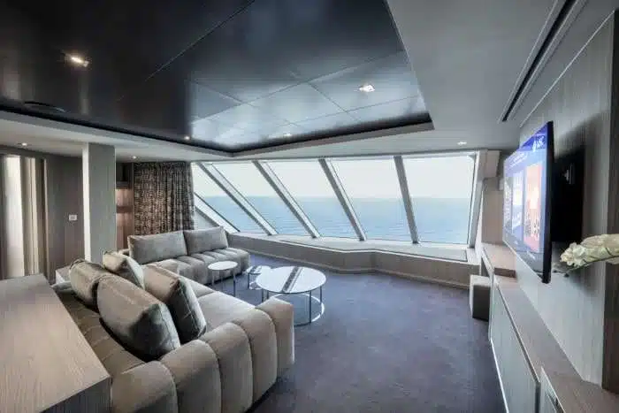 A modern, spacious cruise ship suite with a large angled window offering an expansive ocean view. The room features a plush gray sectional sofa, minimalist furniture, and a wall-mounted flat-screen television.