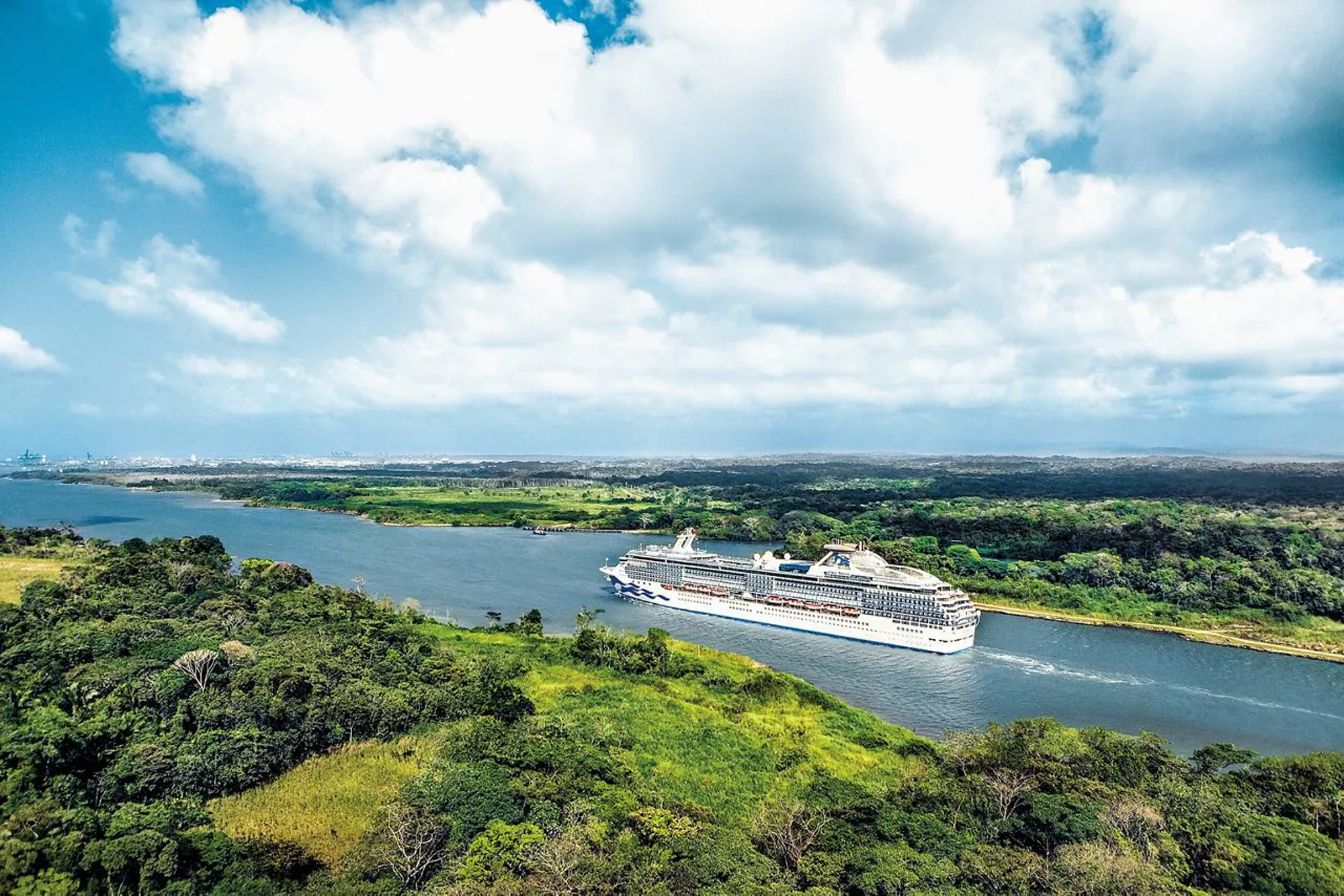 Cruise Ship Navigating Tropical Waterway A large white cruise ship sailing through a lush green landscape with dense forests and blue waters. The ship is framed by verdant coastlines under a bright, cloudy sky.