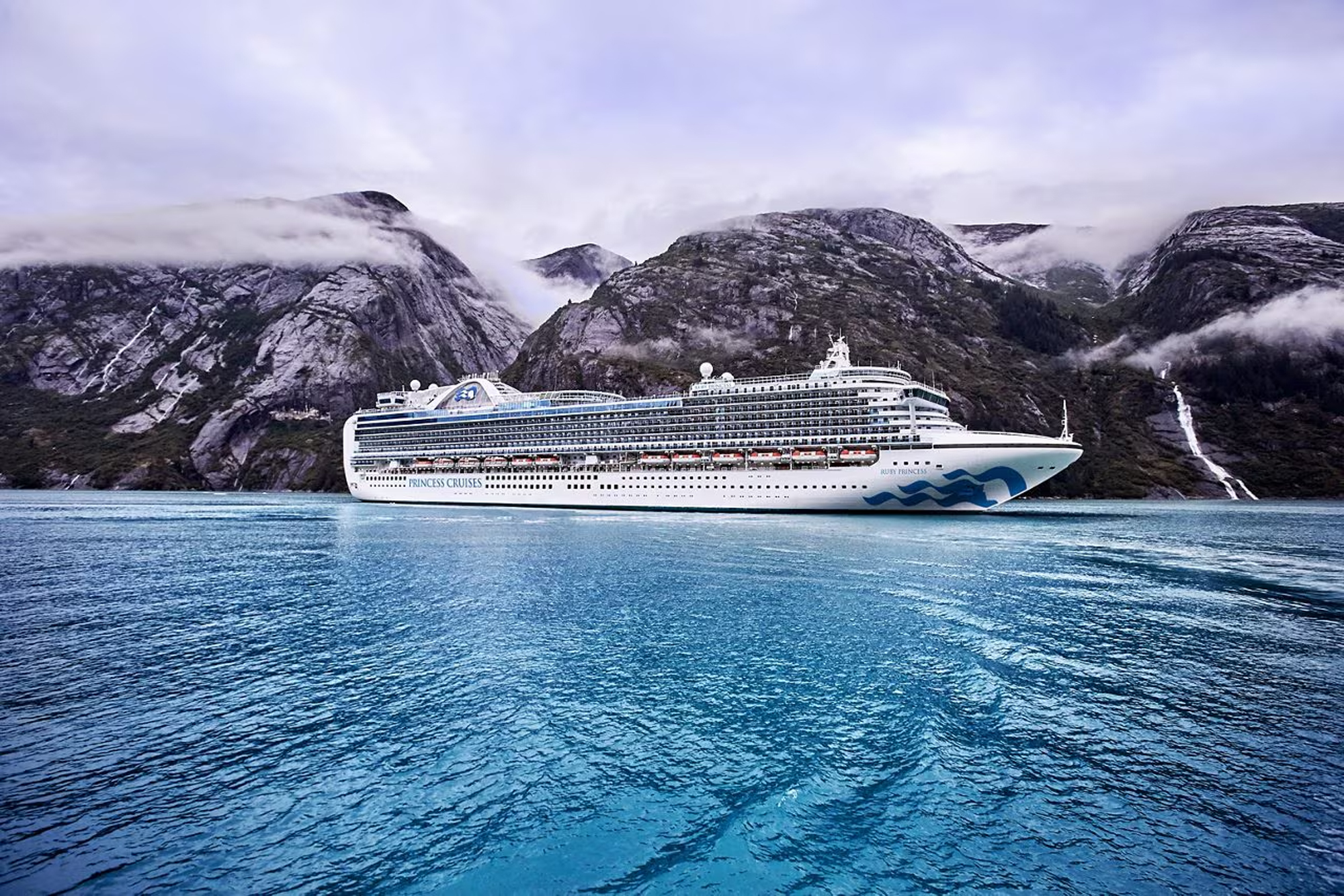 Cruise Ship in Misty Alaskan Fjords A large white Princess Cruises ship sailing through a dramatic fjord landscape with steep rocky mountains and low-hanging clouds. The blue waters reflect the surrounding mountainous terrain.