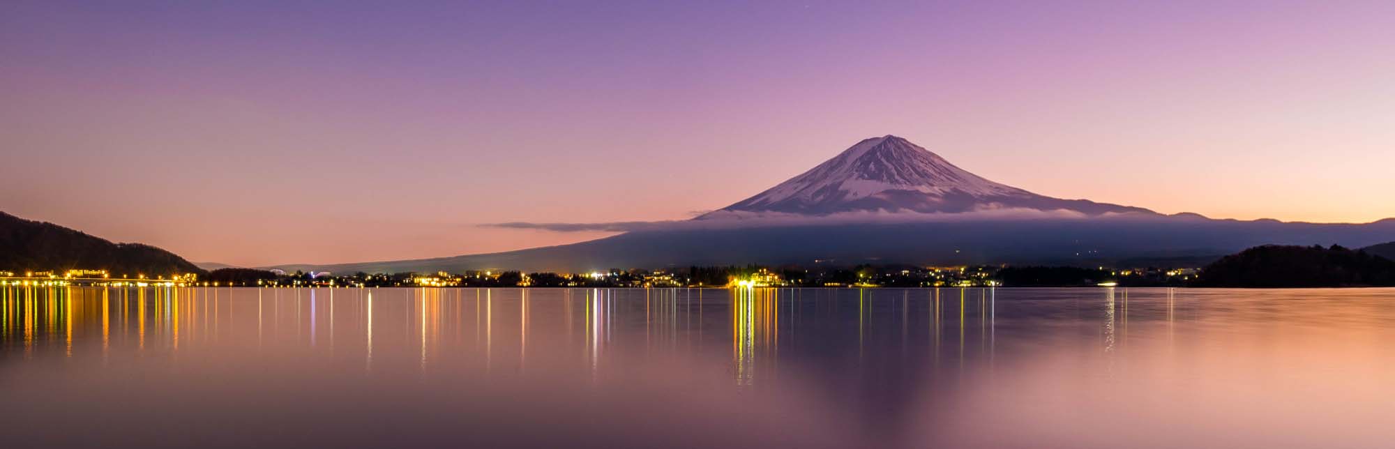 A stunning panoramic view of the iconic Mount Fuji reflecting in calm waters at twilight, with city lights illuminating the shoreline in soft golden and purple hues.