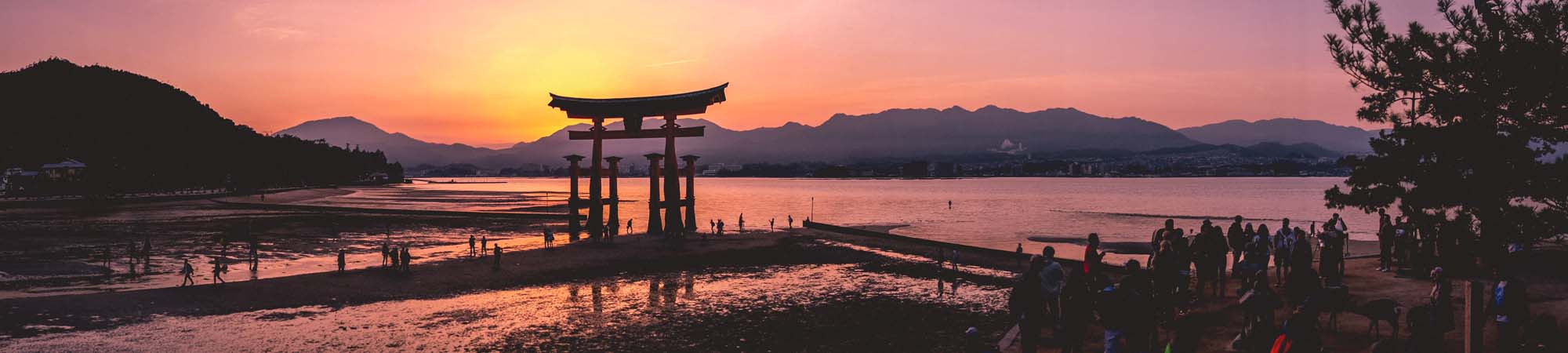 A stunning sunset view of the iconic Itsukushima Shrine's floating torii gate at low tide, with silhouetted visitors and mountains in the background.